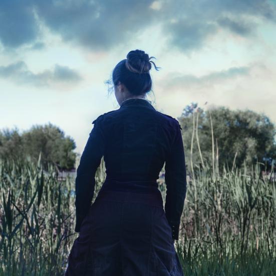 Billedet forestiller en kvinde set fra ryggen. Hun er iført en gammeldags mørk kjole og har sit mørke hår sat op i en knold.