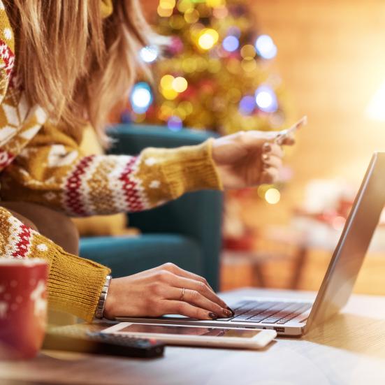 Billedet forestiller en kvinde i en lysebrun julesweater. Hun står bøjet ind over et bord med den ene hånd hvilende på tastaturet på den bærbare computer. 
