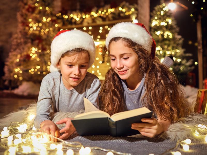 Billedet forestiller to børn der ligger på maven på gulvet. De kigger i en bog. Begge har nissehuer på og i baggrunden kan ses et juletræ.