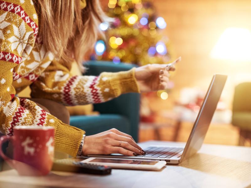 Billedet forestiller en kvinde i en lysebrun julesweater. Hun står bøjet ind over et bord med den ene hånd hvilende på tastaturet på den bærbare computer. 